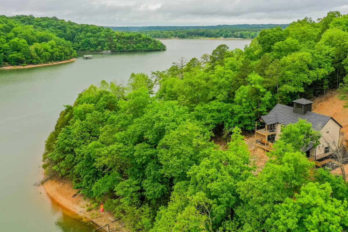 A scenic view showcases a house nestled among lush green trees along the shores of a serene lake. The shoreline curves gently, reflecting the surrounding greenery and clouds in the calm water, creating a peaceful outdoor environment.
