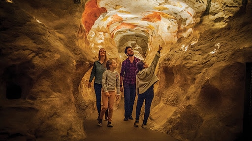A group of four people is seen exploring a cavernous space with textured walls resembling natural rock formations. Soft lighting highlights the unique patterns in the stone, and one individual is pointing towards colorful markings on the ceiling.