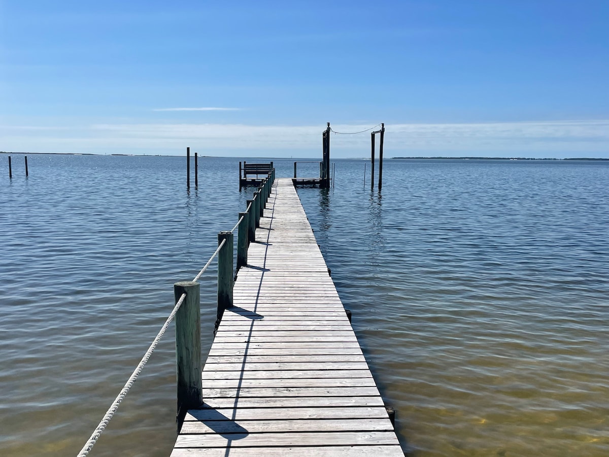 A wooden dock extends over calm waters, leading towards the horizon. The clear blue sky complements the tranquil surface of the water, with a few posts visible along the sides of the dock. The scene evokes a sense of peaceful outdoor exploration.