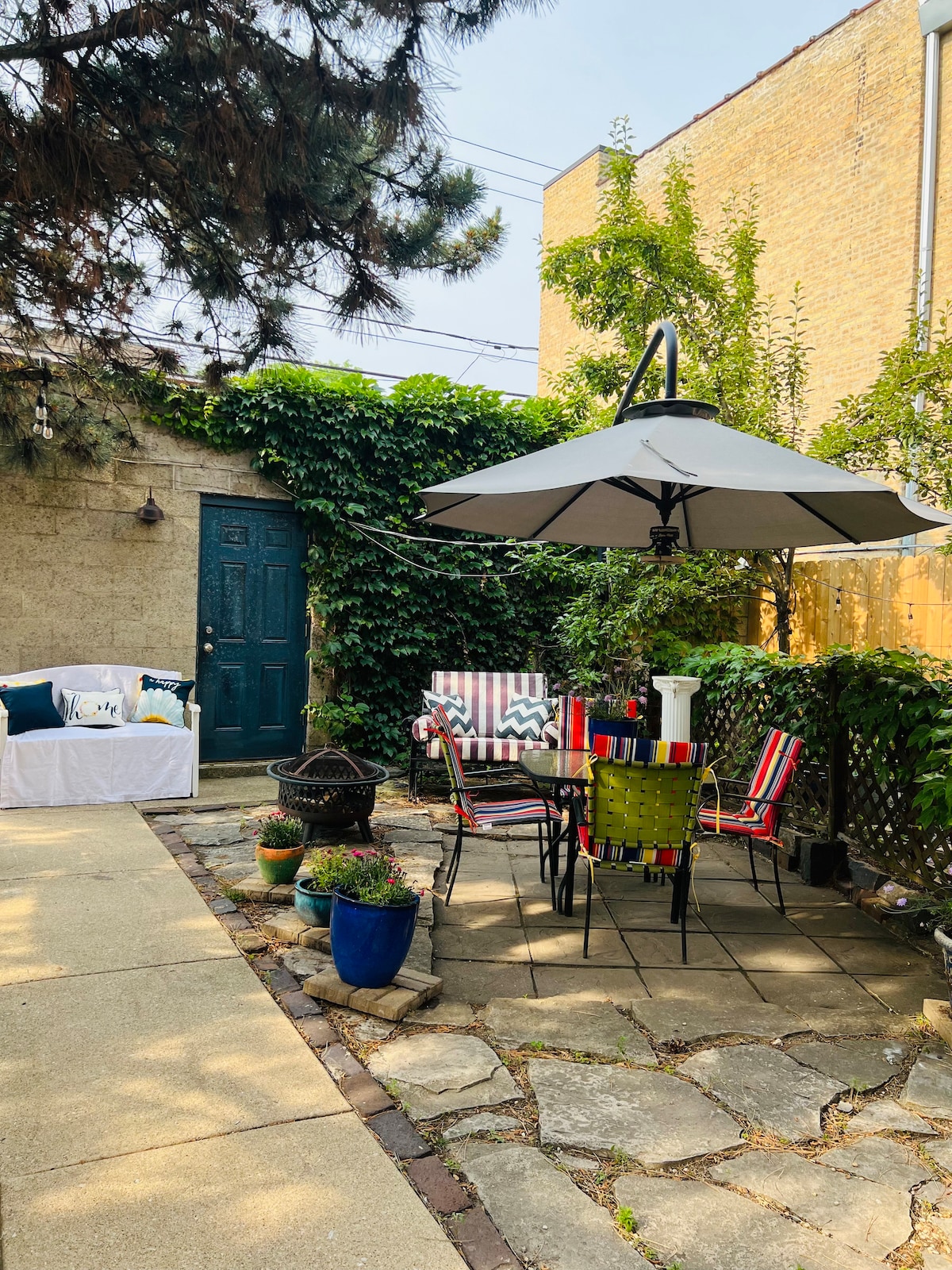 A charming outdoor patio is featured, with a circular table surrounded by colorful chairs. A large umbrella provides shade over the seating area. Various potted plants and a decorative fire pit enhance the space, while a stone pathway leads to an entrance door framed by lush greenery.