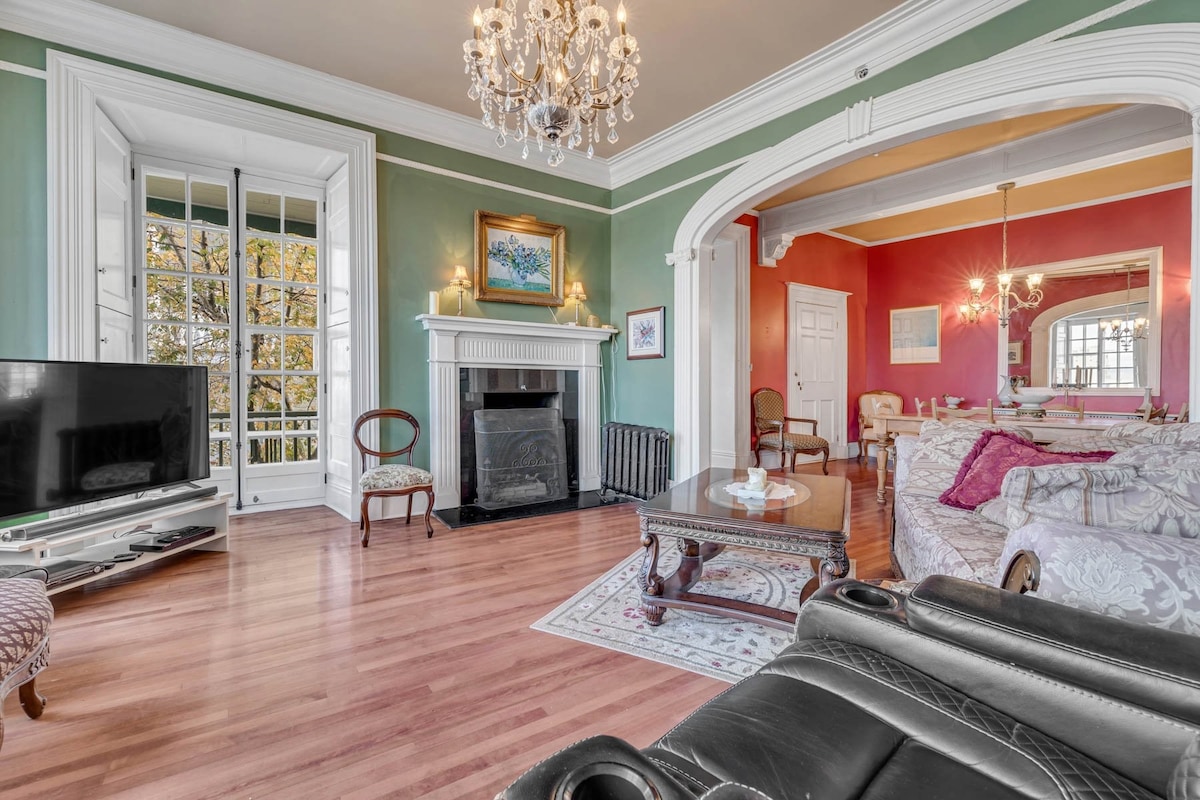 A spacious living area features large windows allowing natural light to fill the room. Elegant furnishings, including antique chairs and a decorative table, surround a traditional fireplace. A chandelier hangs from the ceiling, complementing the unique color palette of the walls.