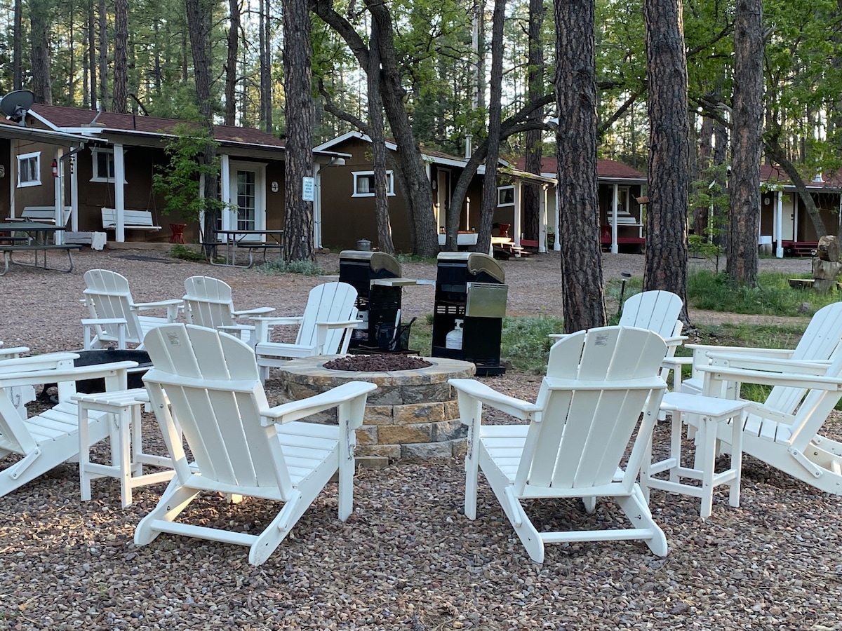 A spacious outdoor area features multiple white Adirondack chairs arranged around a circular stone fire pit. Two propane grills are positioned alongside the fire pit, nestled among tall trees and gravel. Several cabins are visible in the background, surrounded by nature.