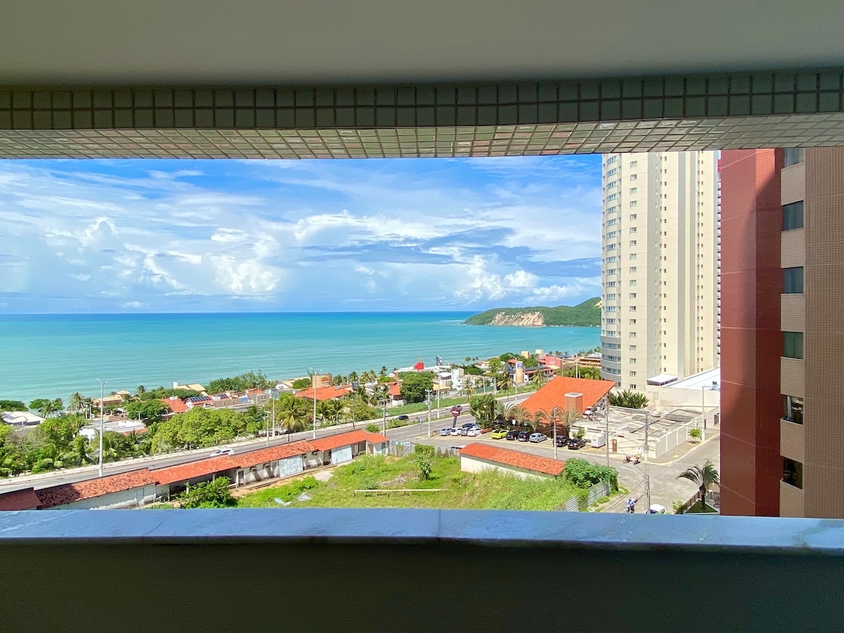 A seascape view is framed by a large window, showcasing the blue ocean meeting a bright sky with scattered clouds. The coastline reveals a variety of buildings and greenery, offering a sense of tranquility in the vibrant coastal setting.