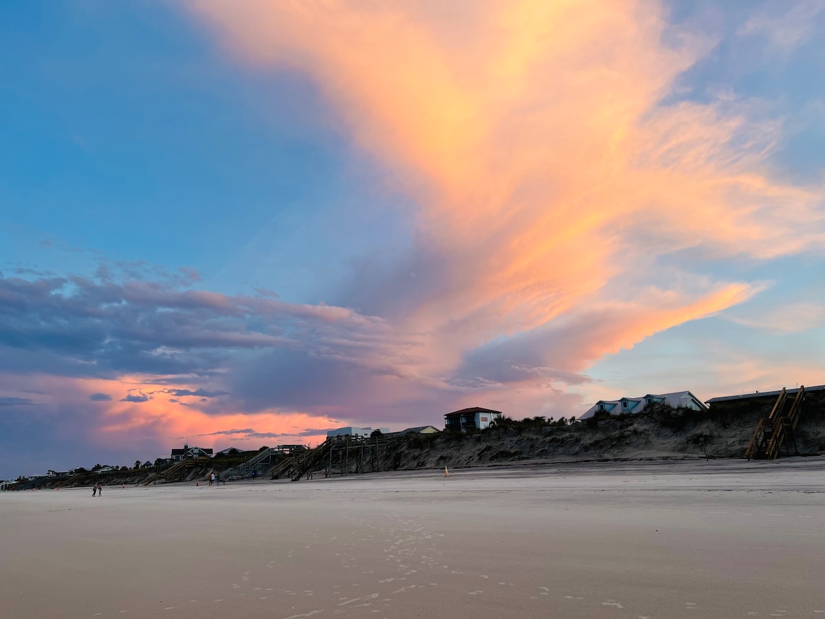 A wide view of a beach at sunset showcases soft pink and orange hues in the sky, complemented by a subtle gradient of blue. Silhouetted structures line the beach, with a sandy shoreline leading into the distance. Gentle clouds drift above, creating a serene atmosphere.