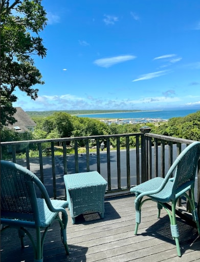 A private balcony is furnished with two teal chairs and a small table, offering a clear view of the harbor and surrounding greenery. The bright blue sky is visible above, with soft clouds scattered throughout.