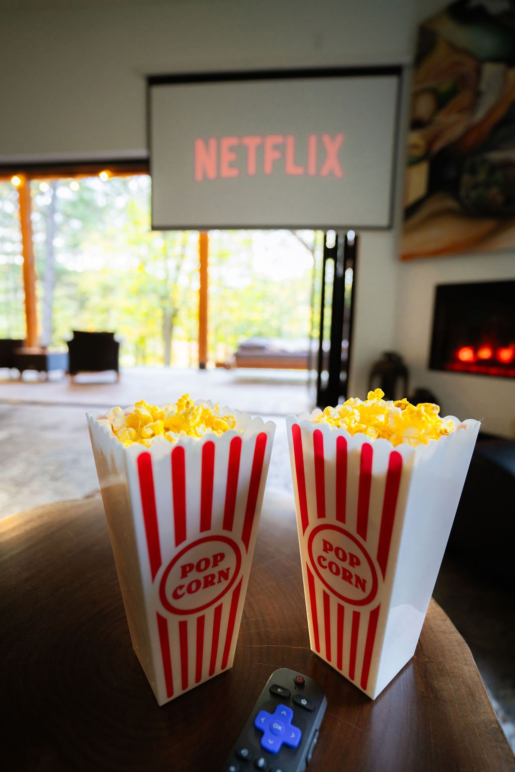 A cozy living space features a large projection screen displaying 'NETFLIX.' Two popcorn boxes are placed on a wooden table, adding a touch of comfort to the inviting setting. Natural light filters through large windows, enhancing the ambiance of the room.