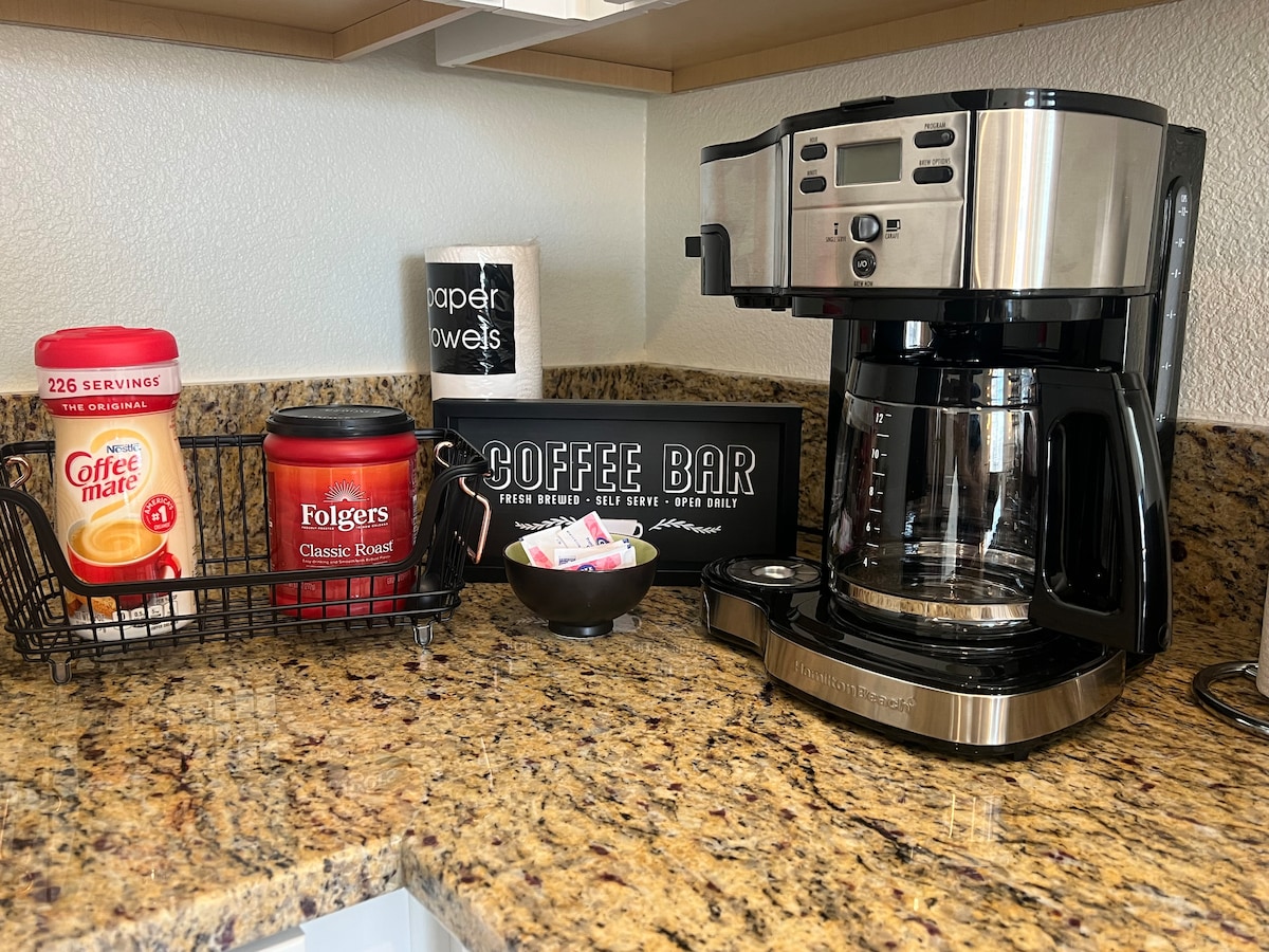 A kitchenette countertop is featured, showcasing a coffee maker beside a basket containing a coffee creamer and a container of classic roast coffee. A decorative sign labeled 'Coffee Bar' sits nearby, adding a functional touch to the well-stocked area.
