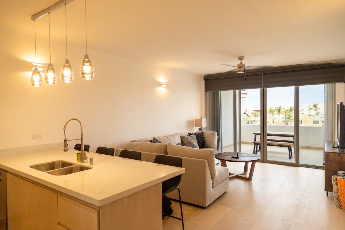 A modern living area features a light-colored sofa paired with a wooden coffee table. The kitchen area includes a sleek countertop with bar stools. Large glass doors provide access to a balcony, illuminated by pendant lights hanging above the kitchen space.