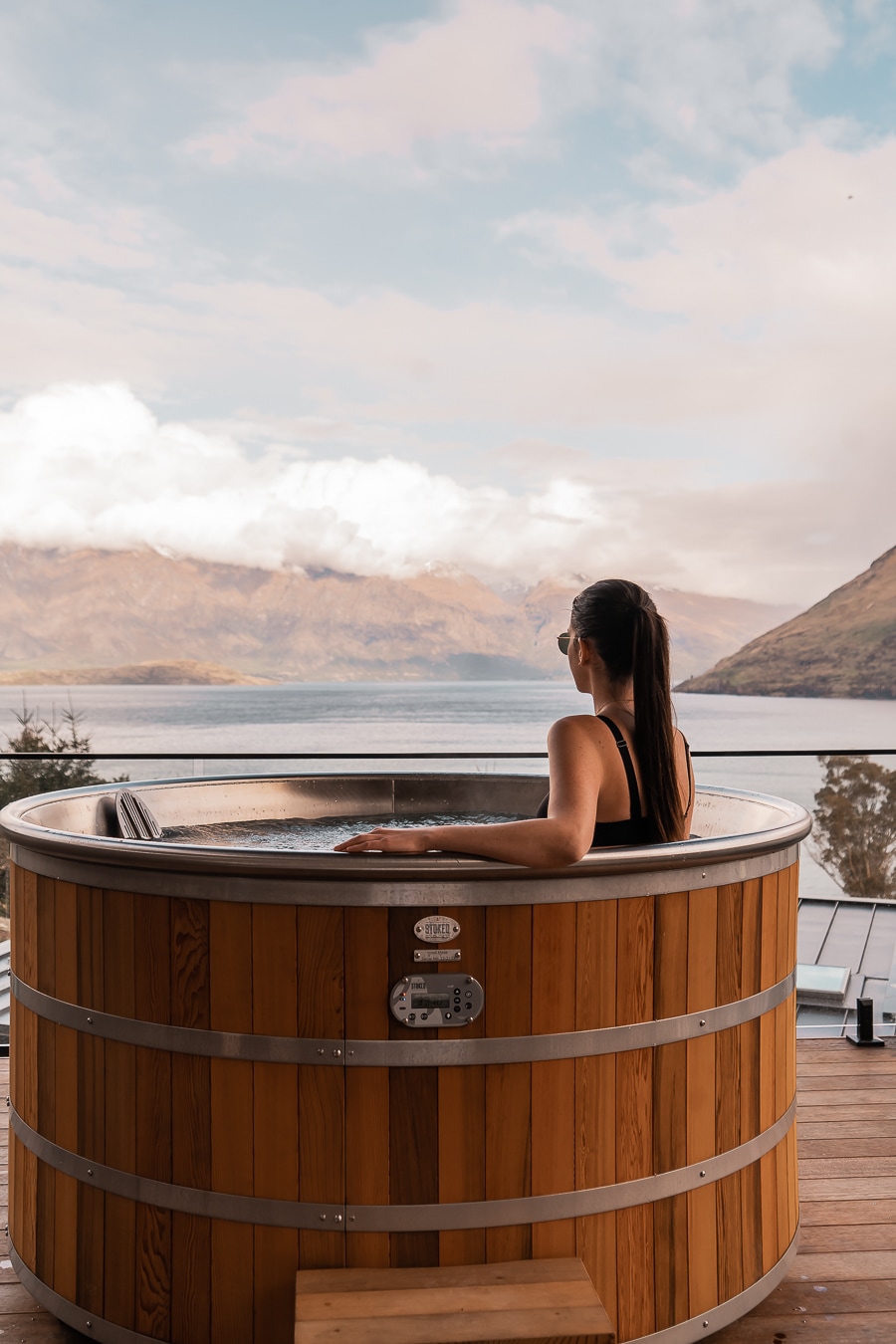 A cedar spa pool is positioned on a deck, providing views of Lake Whakatipu and surrounding mountains. A person sits comfortably in the water, facing the landscape. Soft clouds drift in the sky, creating a serene atmosphere.
