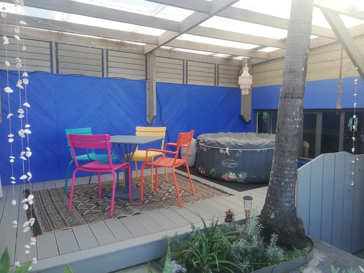 A covered terrace features colorful seating arrangements around a central table, complemented by a patterned rug. A jacuzzi is positioned in one corner, and vibrant blue walls enhance the inviting space. Greenery frames the area, contributing to a relaxed outdoor atmosphere.