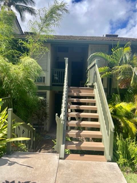 A well-maintained path leads to a set of wooden stairs adorned with a gentle green railing. Lush tropical foliage surrounds the entrance, adding to the serene atmosphere. The inviting stairway ascends toward a covered porch, flanked by palm trees and vibrant plants.