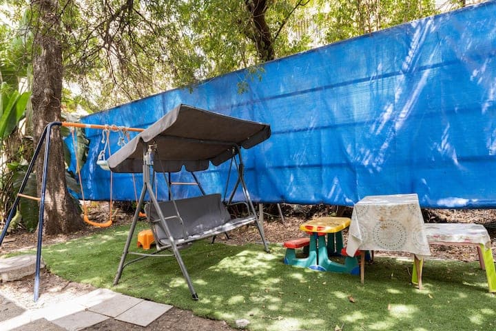 An outdoor area is shown with a swing set and a covered seating area. A small table with multiple colorful chairs is positioned nearby, alongside a grassy surface. A blue tarp provides shelter, framing the space with lush greenery in the background.