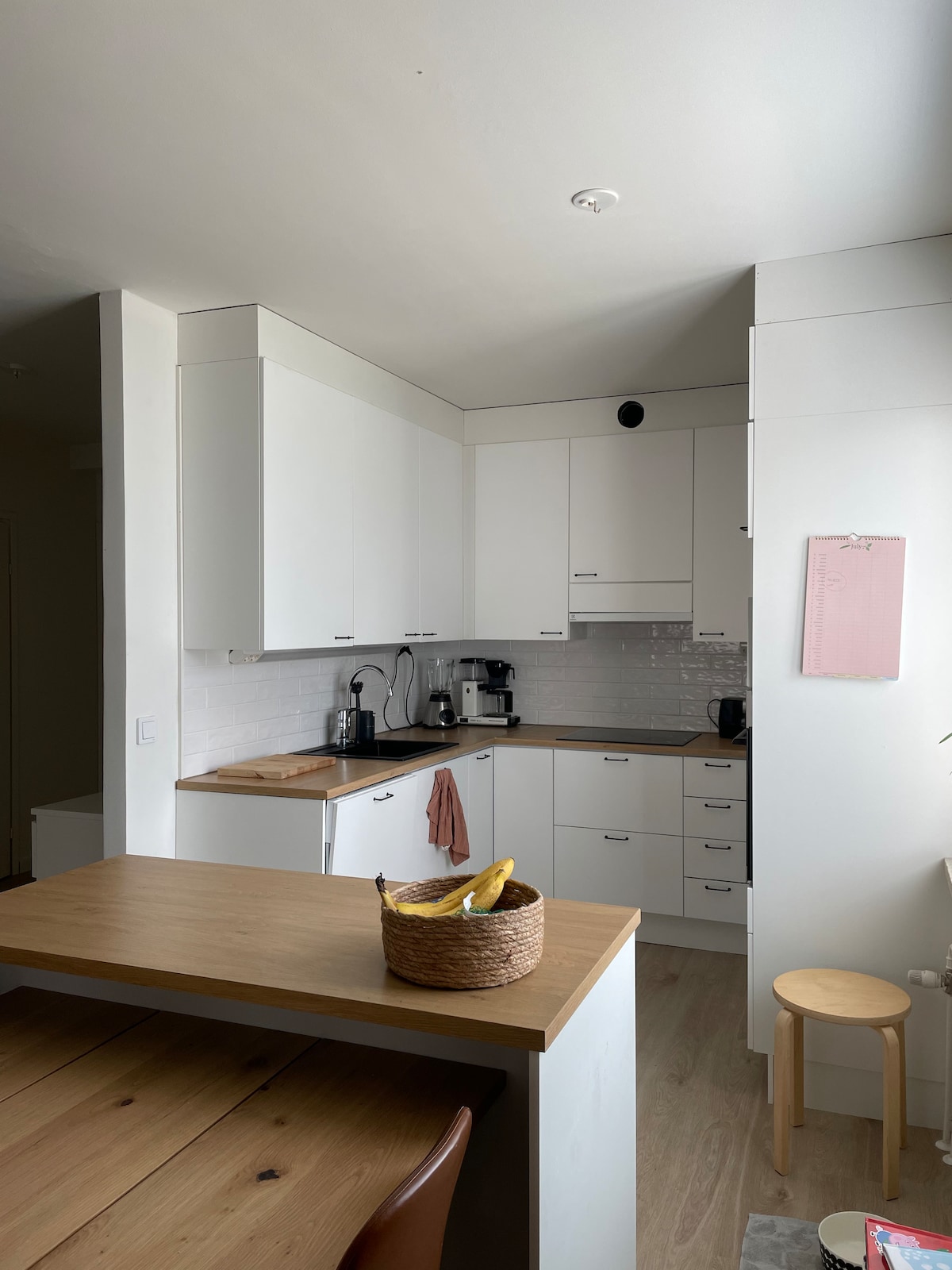 A modern kitchen is equipped with white cabinetry and sleek countertops. Essential appliances, including a coffee maker, are visible. A wooden dining table sits nearby, accompanied by a simple stool and a basket of bananas, adding a touch of casual functionality to the space.