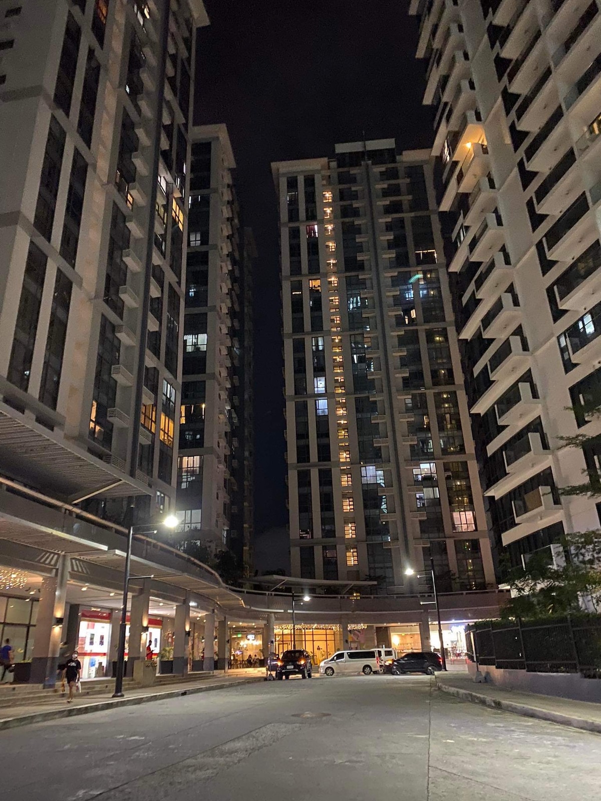Tall residential buildings are illuminated at night, showcasing a mix of glass and concrete facades. Streetlights cast a soft glow on the ground level, where a few pedestrians can be seen walking. The scene conveys a modern urban environment.