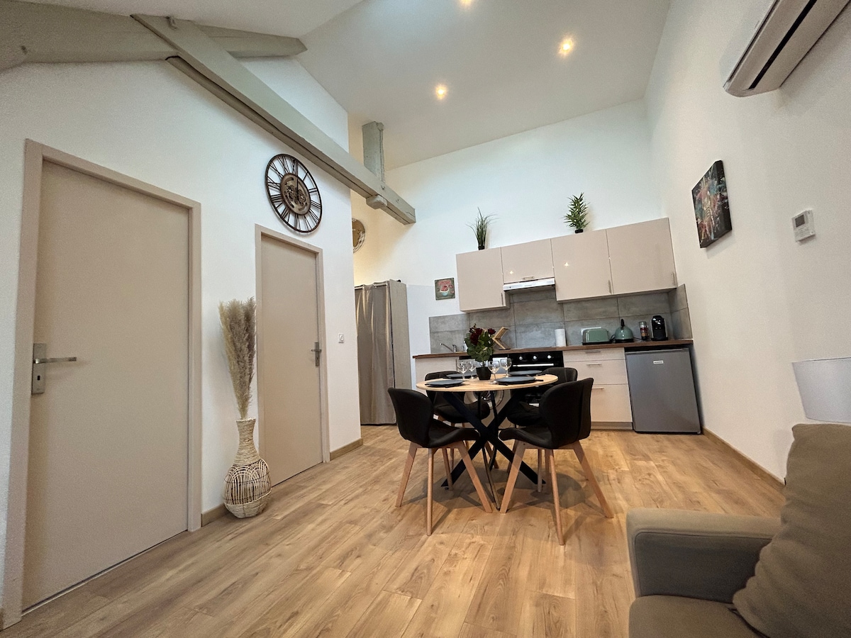 A modern kitchen area is visible, featuring light cabinetry and sleek appliances. A round dining table is set for four, complemented by black chairs. Light wooden flooring extends throughout the space, while decorative plants add touches of greenery to the room.