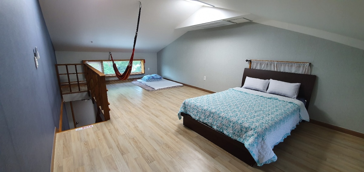 A spacious loft area contains a large bed with a light blue patterned comforter and two pillows. A cozy blue mat is placed on the floor, and a hammock is suspended near a window, allowing for relaxation. Natural light brightens the wooden floors.