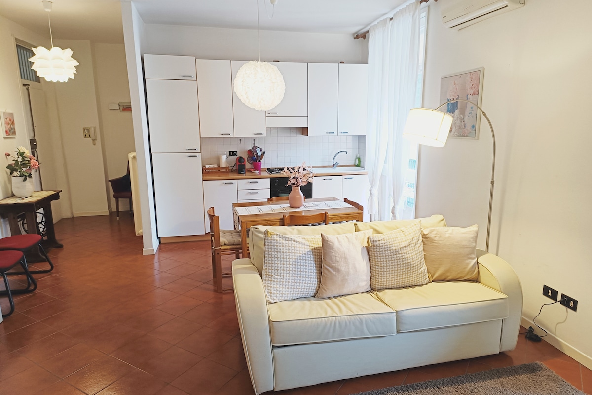 An inviting living space is highlighted, featuring a light-colored sofa adorned with cushions. A wooden dining table is visible, accompanied by chairs in the background. The kitchen area, equipped with white cabinets and appliances, offers a functional layout with natural light filtering through sheer curtains.