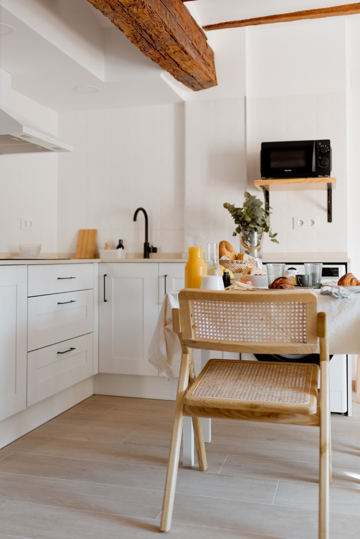 A modern kitchen area is presented with white cabinetry and wooden accents. A small table is set with breakfast items, including pastries and beverages. A microwave and sink are visible, along with a warm wood beam across the ceiling, adding rustic charm.