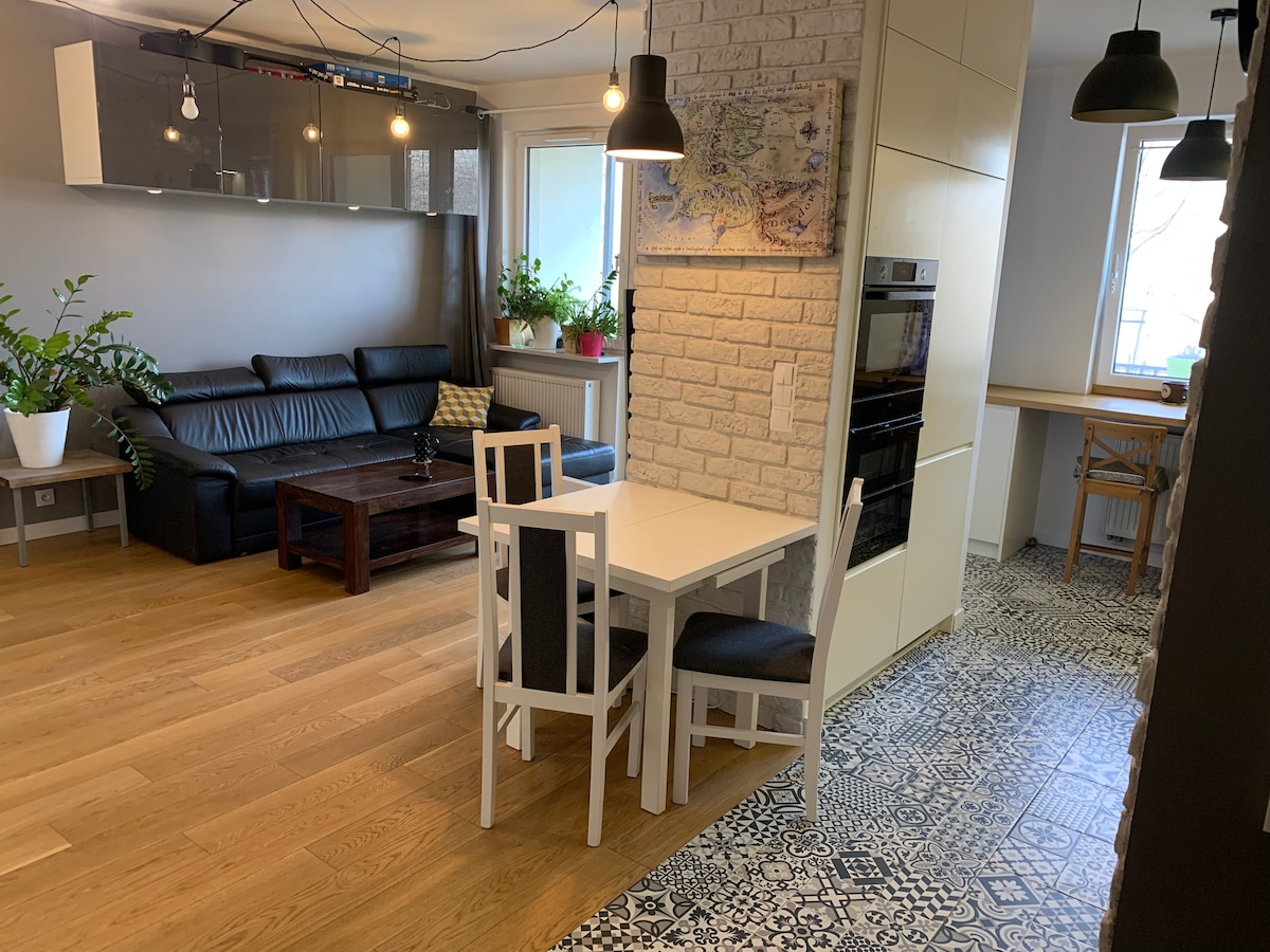 A modern living space is visible, featuring a dark leather sofa against a light brick wall. A dining table with white chairs is positioned beside a sleek kitchen. Natural light filters through large windows, illuminating patterned flooring and a variety of indoor plants.