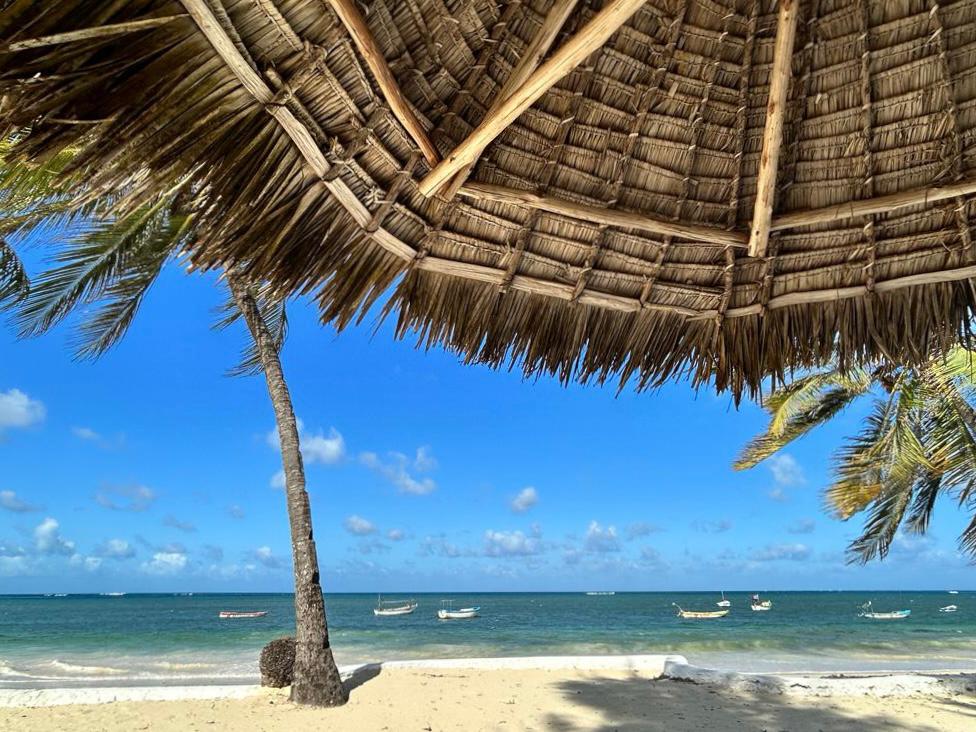 A thatched umbrella provides shade over a sandy beach area. Gentle waves lap at the shore, with a few boats visible on the water in the background. Palm trees sway lightly in the breeze, creating a serene coastal scene.
