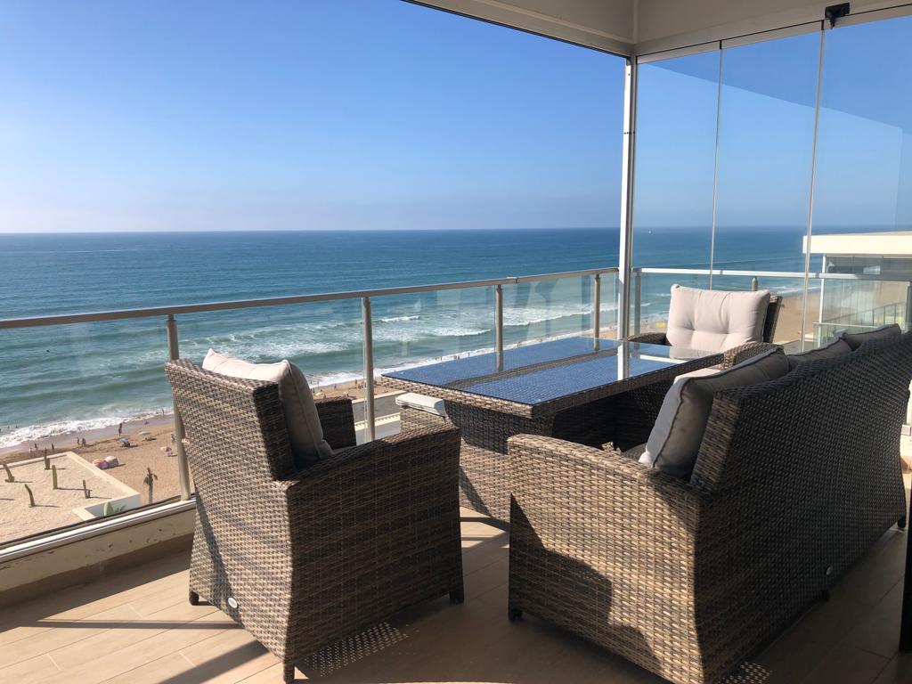 A spacious balcony features comfortable wicker seating with soft cushions, overlooking the Atlantic Ocean. The glass railing allows for unobstructed views of the sandy beach and waves, enhancing the outdoor relaxation space bathed in natural light.