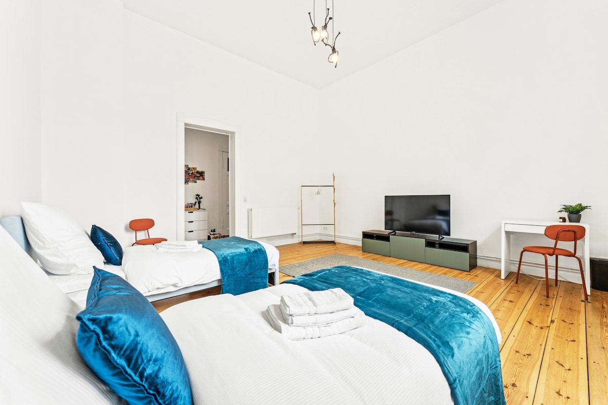 A spacious bedroom features two separate beds with blue throws and neatly arranged towels. A dark television stand with a large screen is positioned across from the beds. A white desk and an orange chair provide a functional workspace. Light wood flooring enhances the room's bright atmosphere.