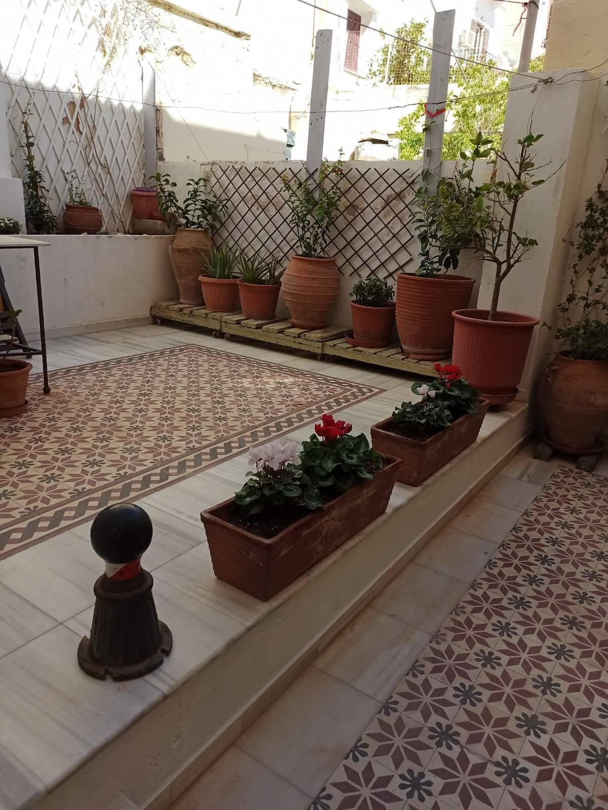 A serene outdoor courtyard features terracotta pots filled with greenery and flowering plants. Decorative tiles cover the ground, enhancing the tranquil atmosphere. Simple seating arrangements are positioned against the wall, providing a peaceful spot to relax in a shaded environment.