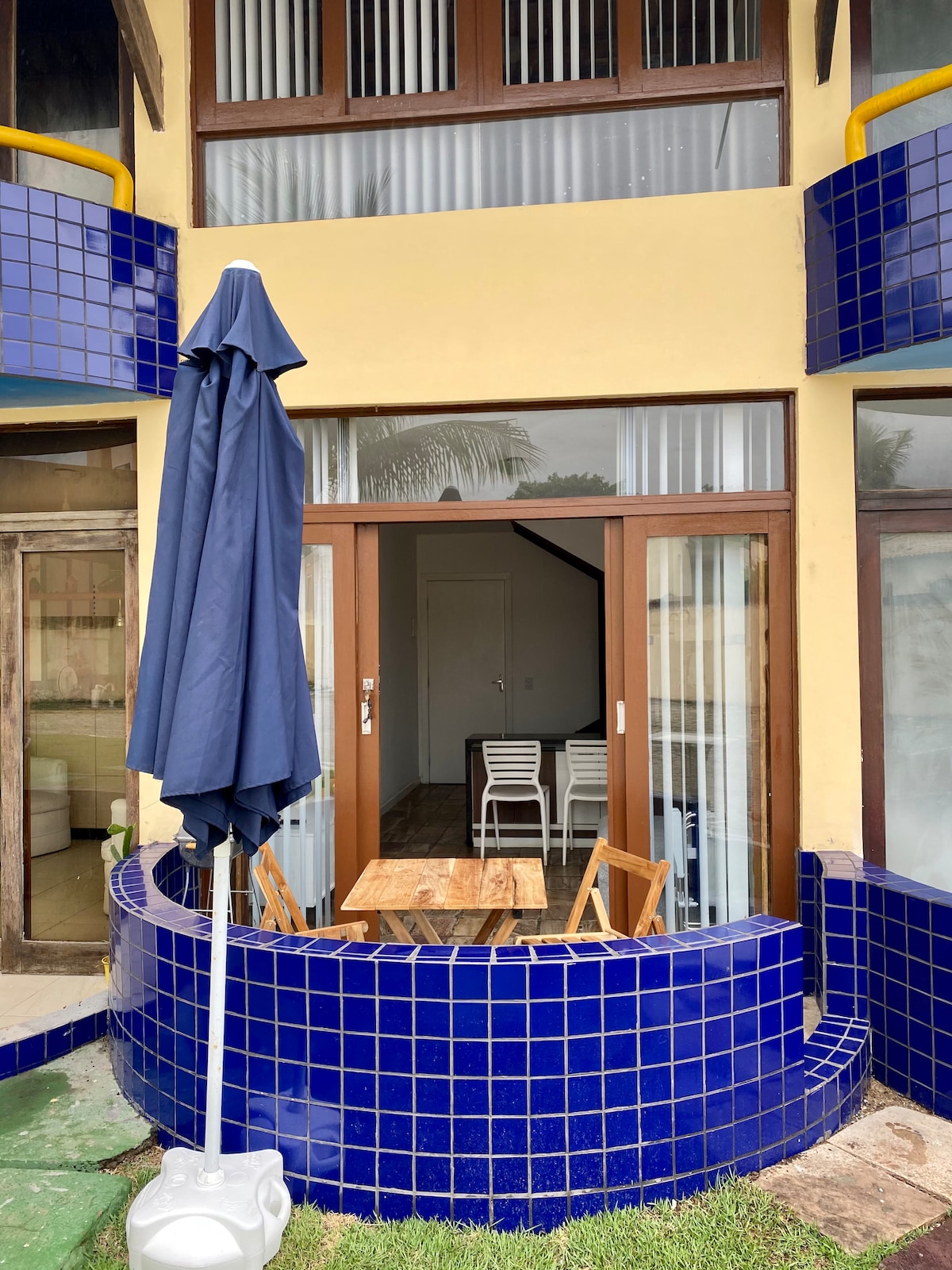 An outdoor patio area is featured, showcasing a wooden table surrounded by two chairs. A large blue umbrella provides shade, while blue tiled walls enhance the space. Glass doors lead into the interior, inviting natural light into the room.