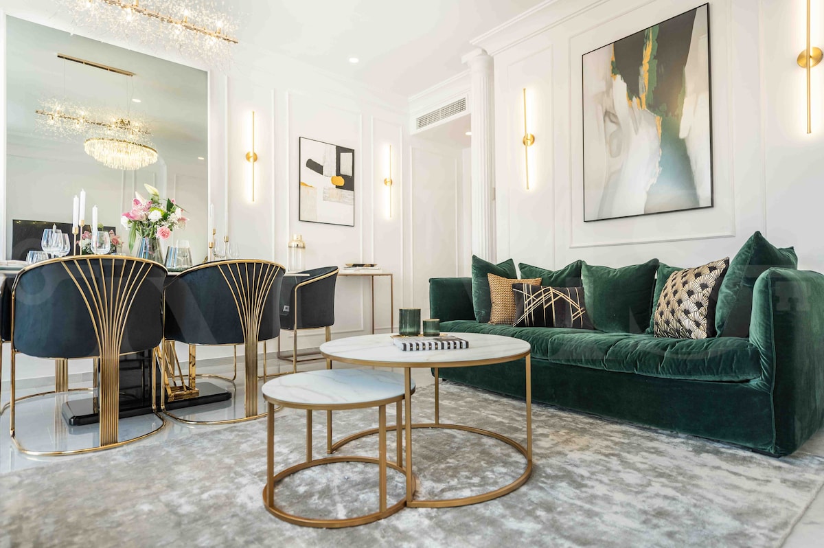The living area is furnished with a plush green sofa and two gold-accented coffee tables atop a soft rug. A dining area features elegant black chairs complemented by a modern chandelier. Wall art and decorative lighting enhance the sophisticated ambiance.
