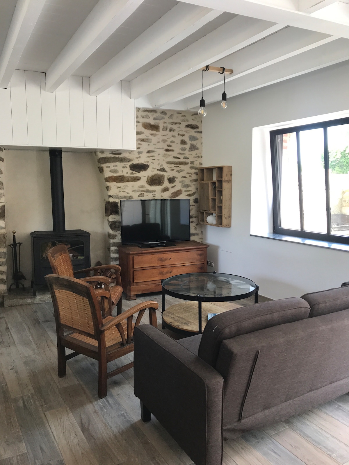 A living area features a wooden ceiling with exposed beams and a stone wall. A modern glass coffee table is positioned in front of a comfortable gray sofa and wicker chairs. A flat-screen TV is mounted on the wall near a classic wood stove, providing a cozy atmosphere.