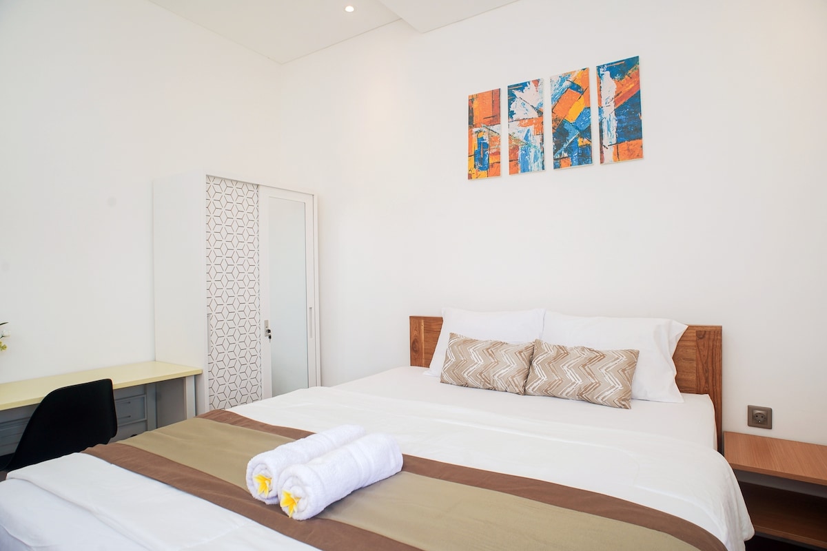 A well-arranged studio room features a spacious bed with decorative pillows and a neutral-toned blanket. A simple wardrobe is positioned beside the bed. Above, colorful artwork enhances the bright white walls, while natural light illuminates the space from an unseen window.