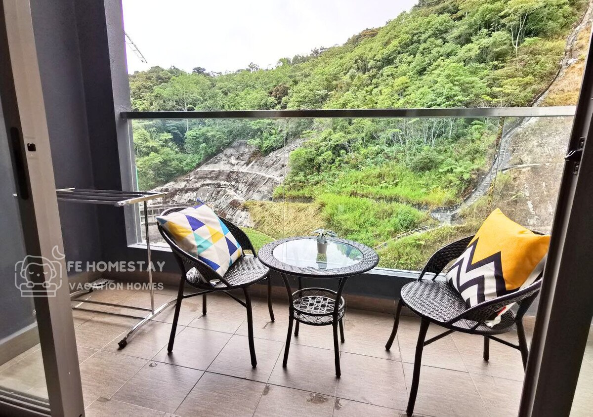 A small balcony features two black rattan chairs with colorful cushions and a round glass table. Lush greenery can be seen in the background, offering a serene view of the hills.