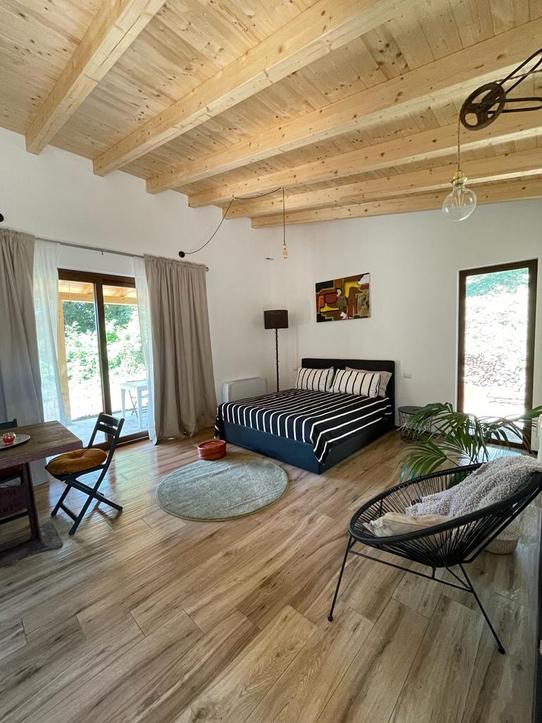 An inviting interior features wooden beams overhead and a spacious bed adorned with black and white striped bedding. Natural light enters through glass doors that lead outside. A small dining area with a wooden table and chairs is visible, accompanied by a cozy area rug.