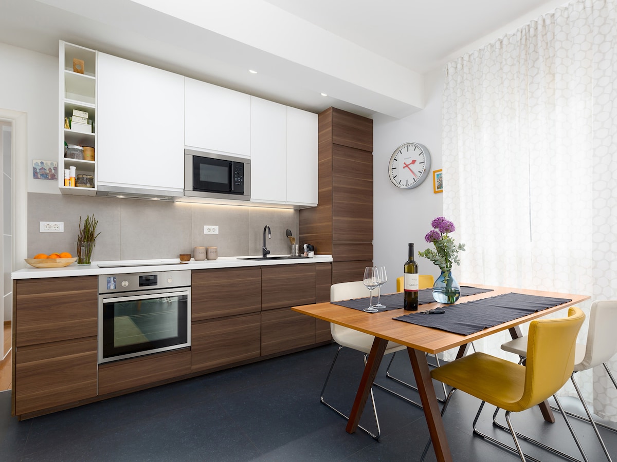 The kitchen area showcases modern cabinetry with a blend of dark wood and white finishes. A dining table is set for two, accompanied by colorful chairs. A window with sheer curtains allows natural light, while kitchen appliances are neatly arranged, presenting opportunities for cooking and dining.
