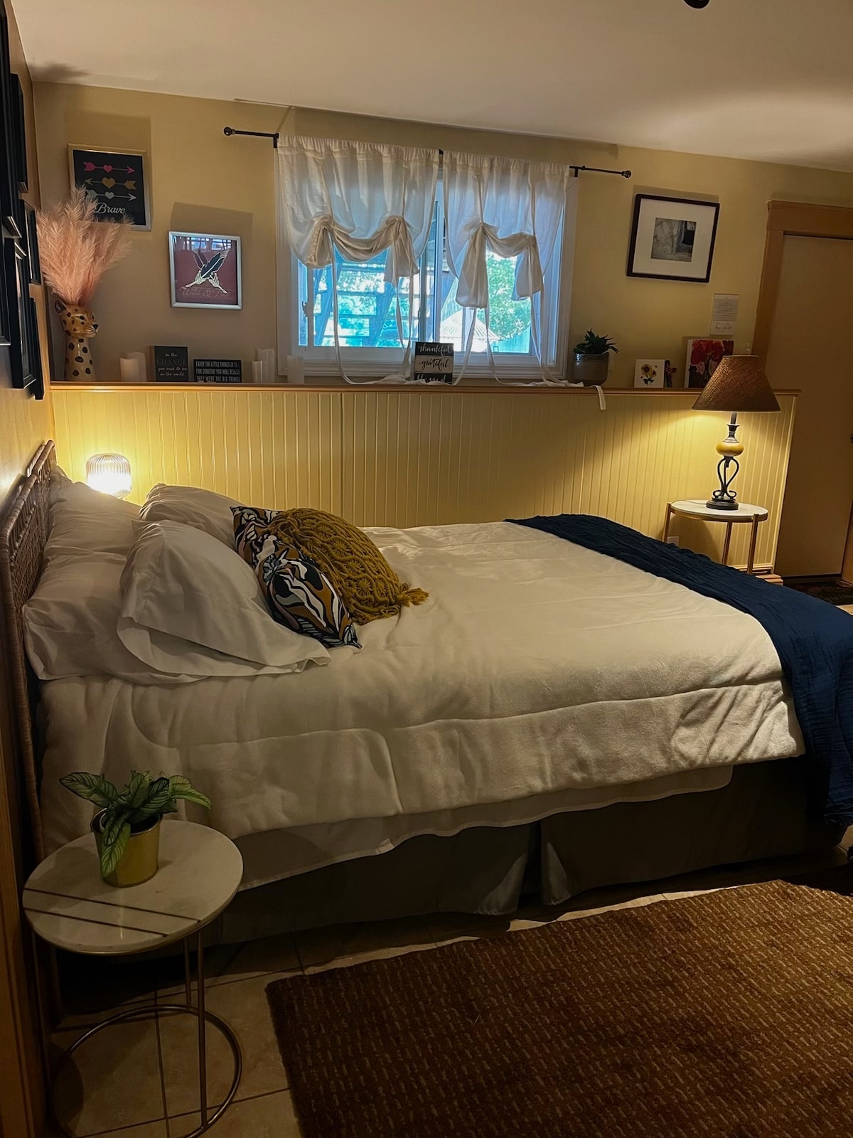 A well-lit sleeping area features a spacious bed with a white comforter and decorative pillows. A small bedside table holds a lamp. Natural light enters through a window dressed with sheer curtains, illuminating the surrounding decor, including framed artwork and personal touches on the walls.