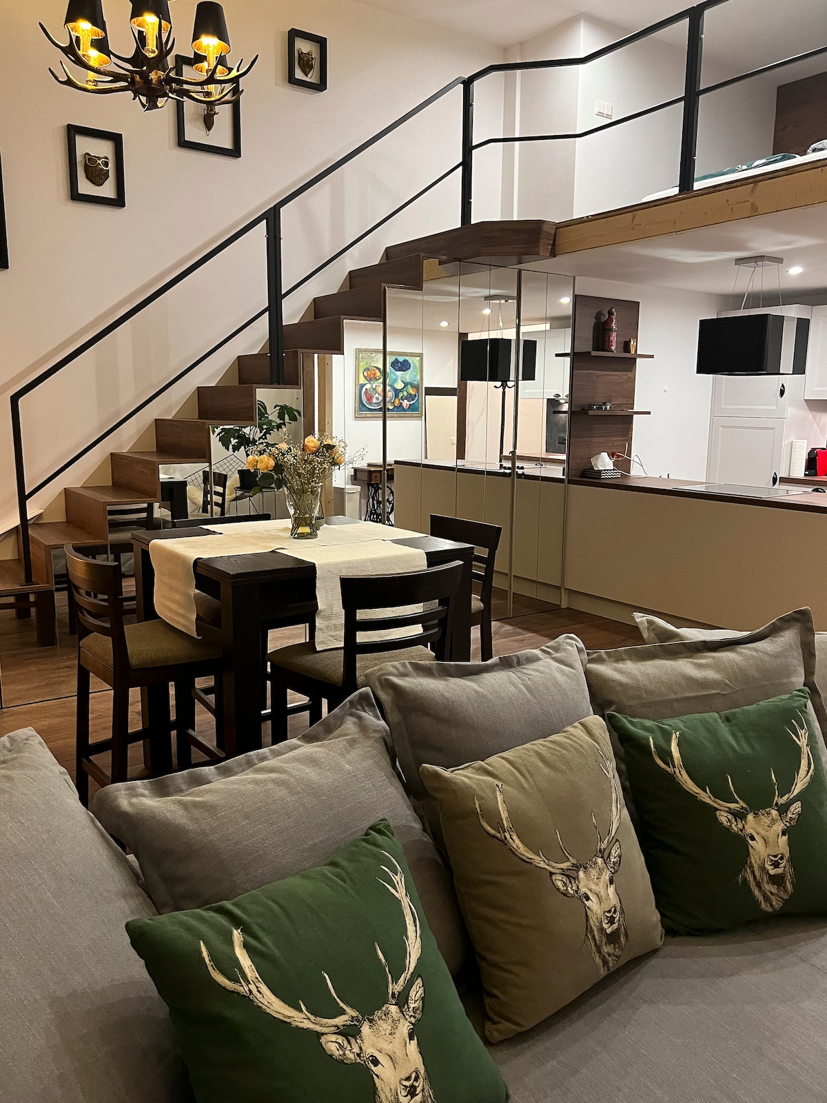 The open living space features a cozy seating area with grey sofas adorned with decorative pillows. A dining table is set for four, accompanied by a view of the modern kitchen with sleek cabinetry and glass panels. A staircase leads to the loft area, enhancing the layout.