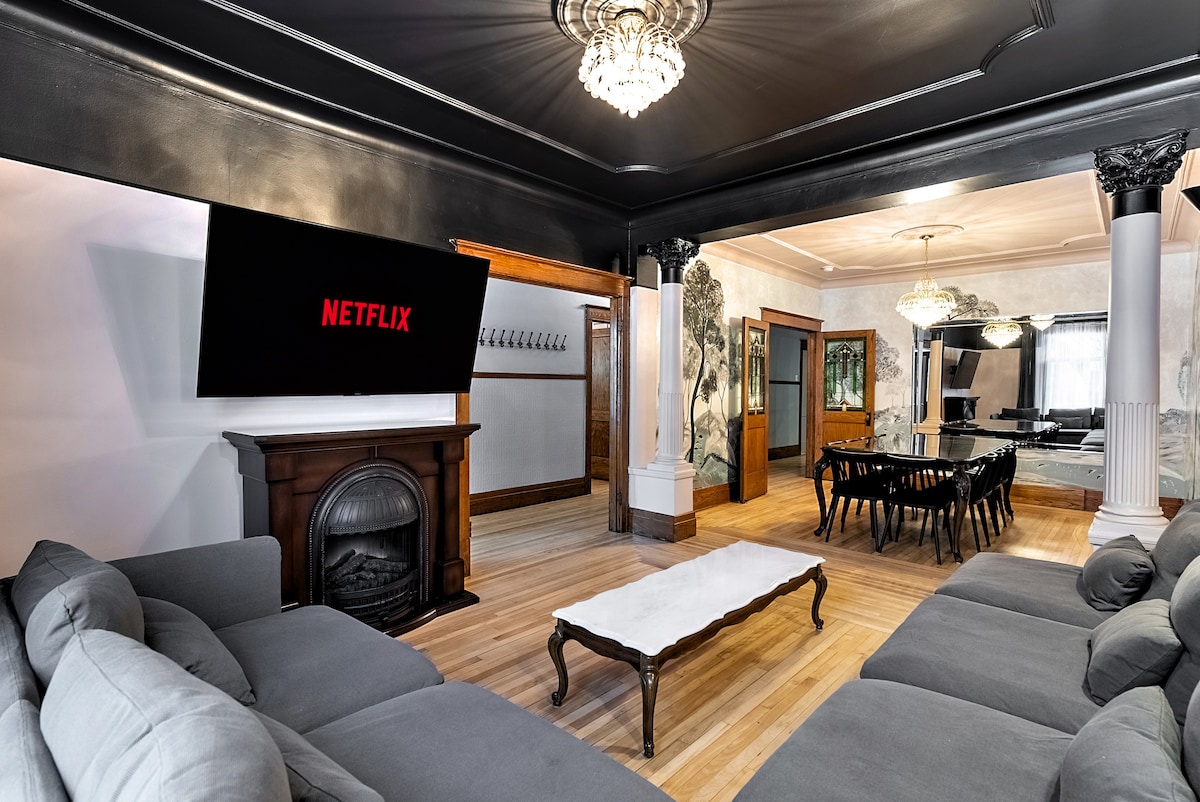 A modern living room features a spacious sectional sofa arranged around a coffee table. A large smart TV displays the Netflix logo, while an elegant electric fireplace adds warmth to the space. The adjoining dining area showcases a long wooden dining table and decorative elements.