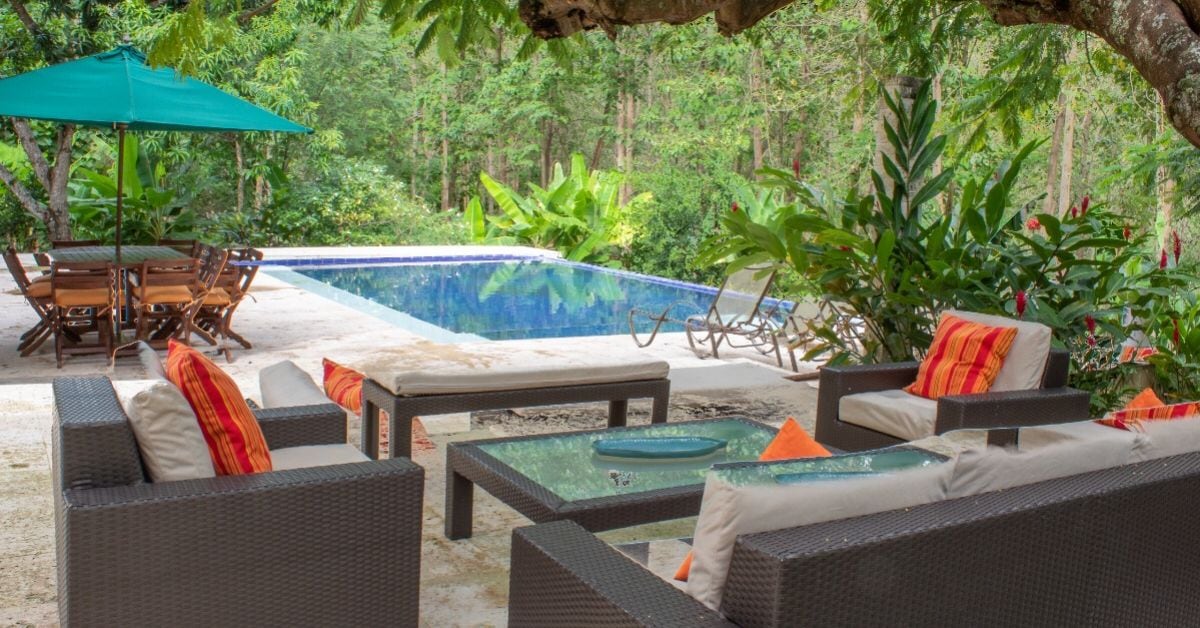 A shaded outdoor area features a seating arrangement with comfortable sofas and colorful cushions. A dining table with wooden chairs is visible, accompanied by a vibrant green umbrella. In the background, a pool is surrounded by lush vegetation, contributing to a serene environment.