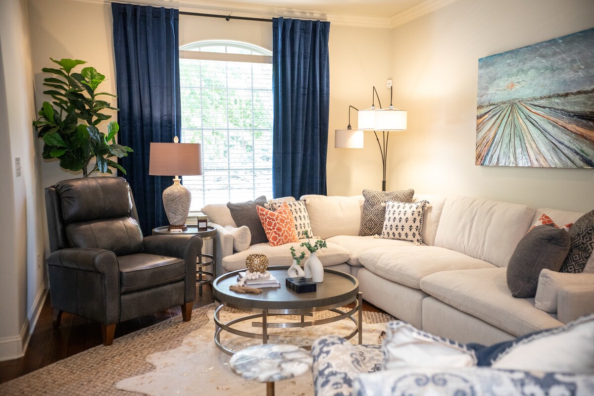 A well-appointed living area features a spacious sectional sofa adorned with decorative pillows. A round coffee table sits at the center, complemented by a stylish lamp and a large window with navy drapes. A piece of abstract art is displayed on the wall.