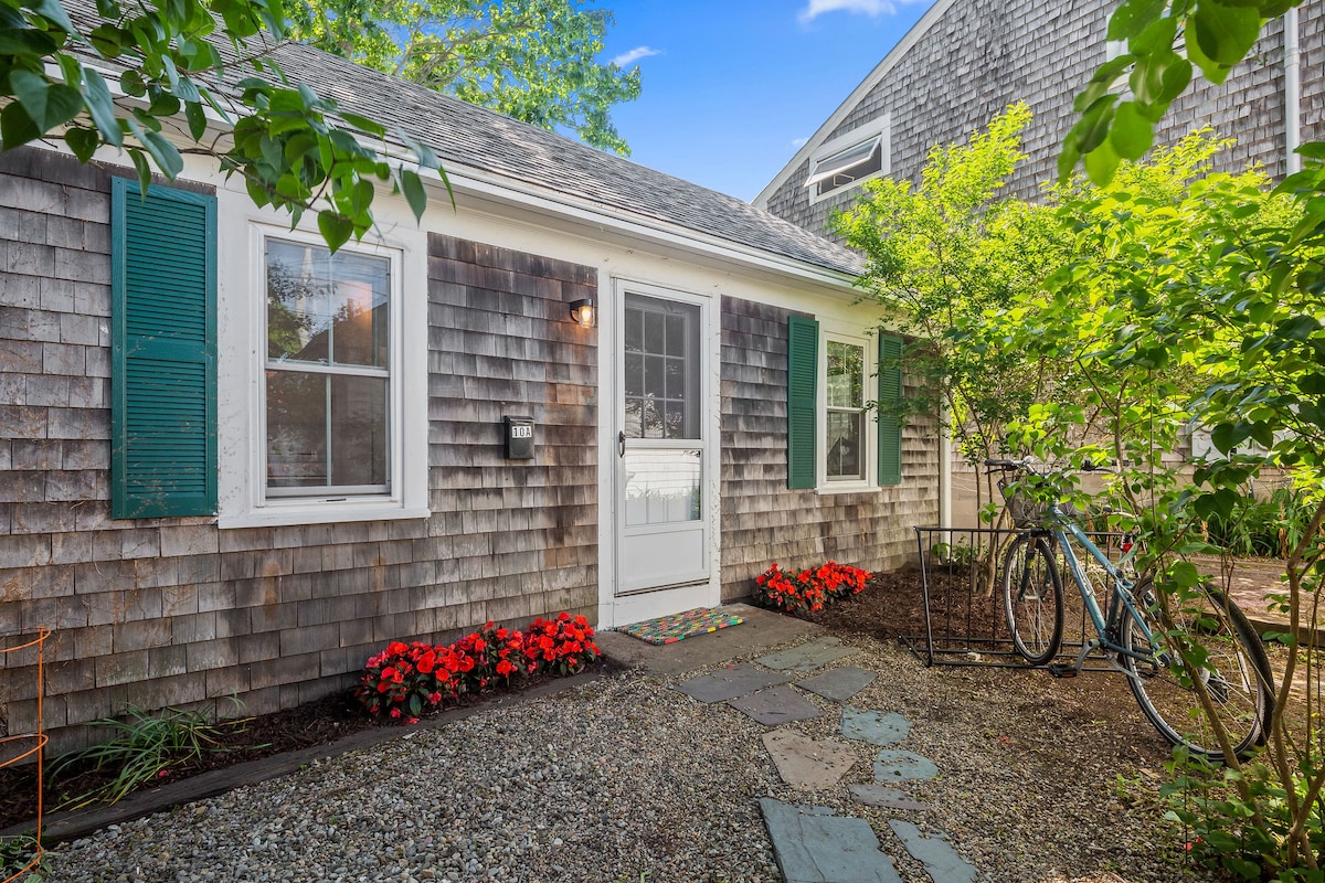 A charming cottage exterior features natural wood shingles and vibrant green shutters. A welcoming white door is flanked by two windows, with blooming red flowers along the gravel pathway. Two bicycles are parked beside a tasteful landscaping of shrubs and trees, enhancing the inviting entryway.