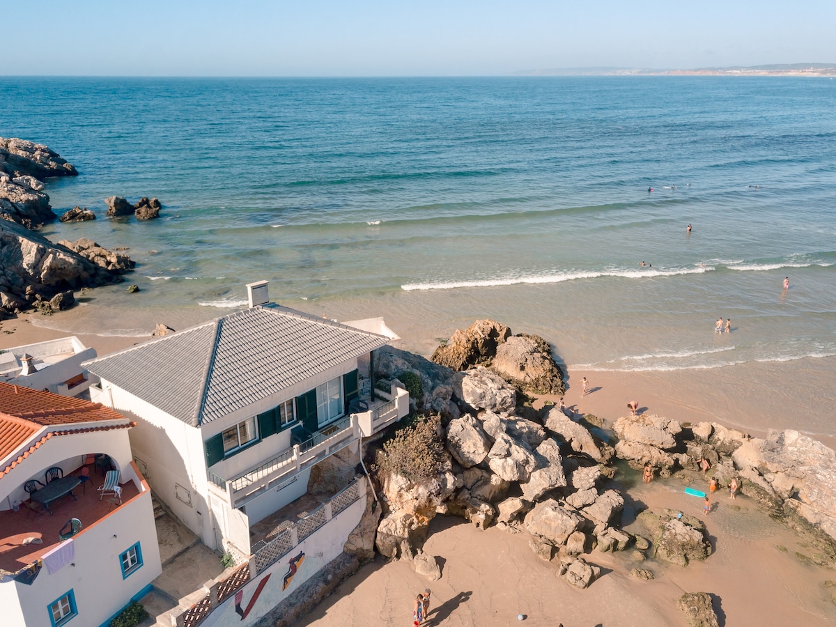 A coastal house is viewed from above, situated near the shoreline. Gentle waves lap at the sandy beach, with people enjoying the water. The structure features a sloped roof, surrounded by rocks and sandy terrain, blending naturally into the coastal landscape.