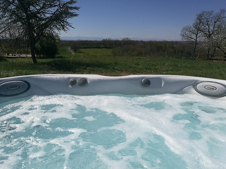 A spacious outdoor jacuzzi is filled with bubbles, set against a backdrop of lush green fields and distant mountains. Clear blue skies above enhance the serene surroundings, creating a tranquil retreat.