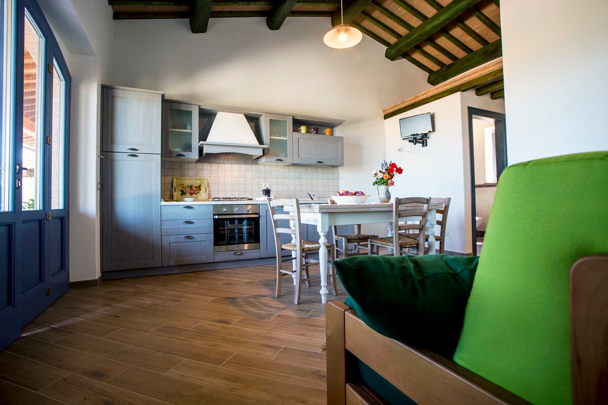 A modern kitchen area is connected to a dining space with a wooden table surrounded by six light-colored chairs. The kitchen features gray cabinetry and stainless steel appliances. Natural light floods the room through large windows, highlighting wooden beams on the ceiling.