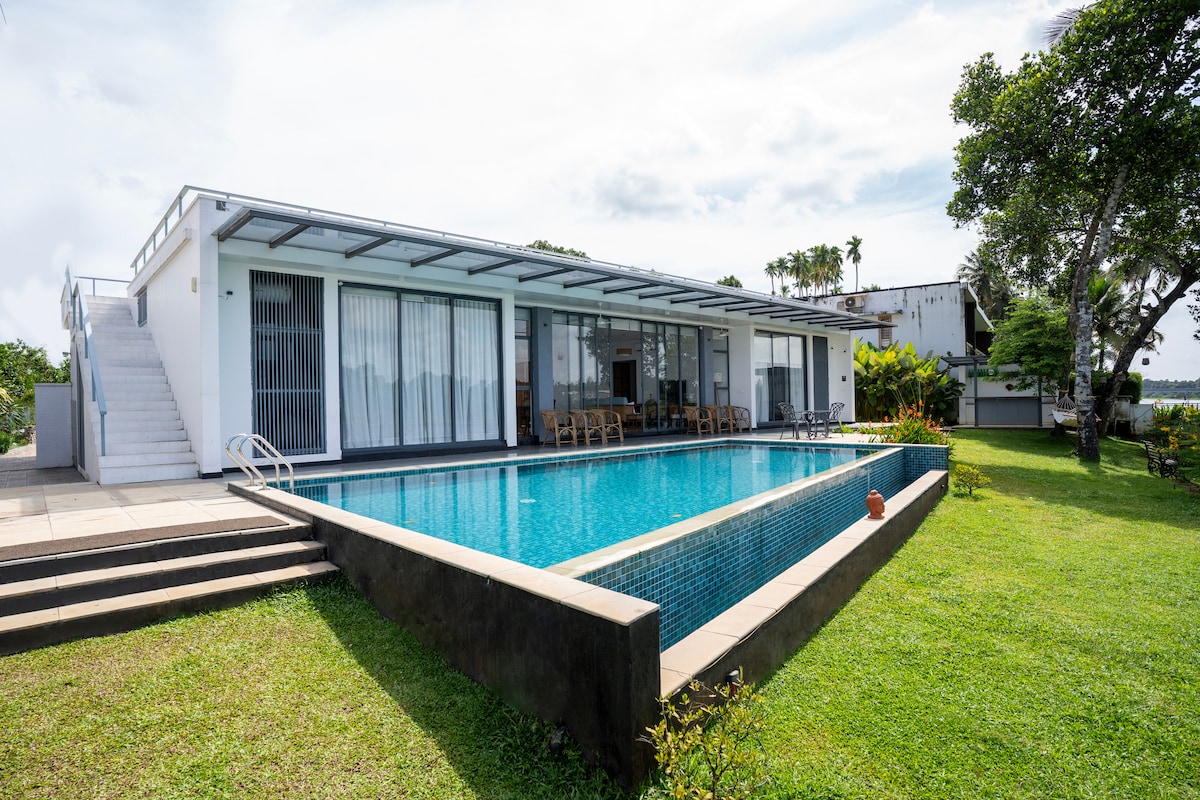 A contemporary villa is presented, featuring an inviting infinity pool bordered by a lush green lawn. Large glass windows reflect natural light, and a spacious deck is accessible from the living area. Steps lead down to the pool, enhancing the outdoor experience.