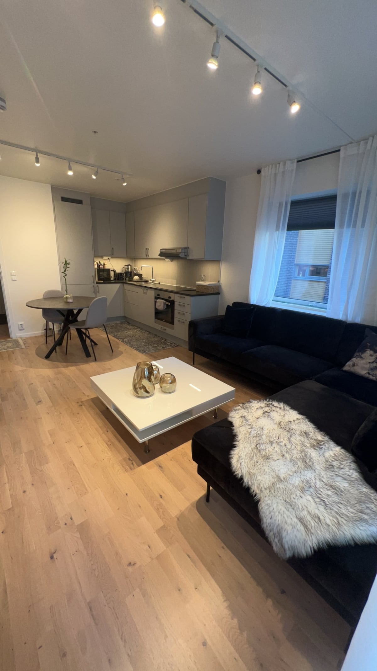 A modern living space features a dark sectional sofa and a light wood floor. A glass-top coffee table with decorative items is centered in the room. A dining area with a small table and chairs is visible. Soft natural light enters through sheer curtains.