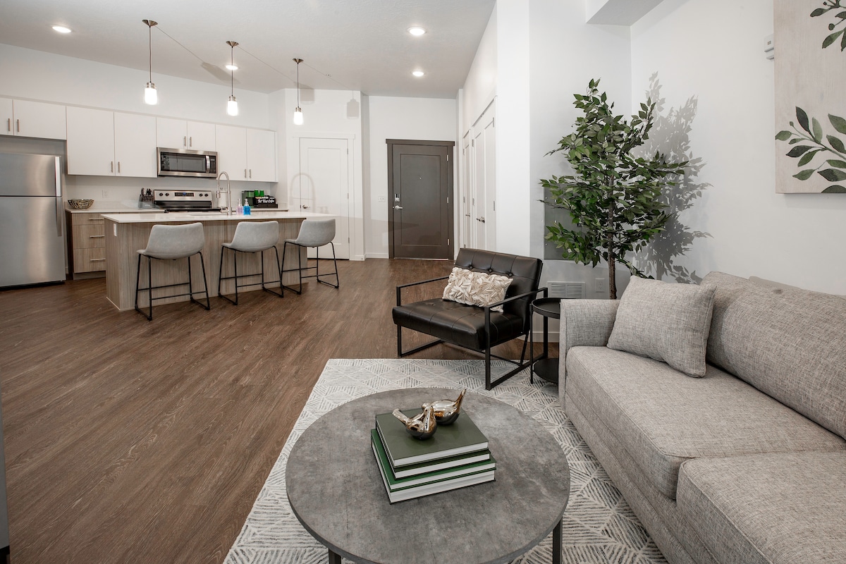 An inviting living area features a cozy gray sofa paired with a black armchair and a decorative coffee table. A modern kitchen is visible in the background, equipped with stainless steel appliances and a spacious island with seating. Natural light floods the space through bright, open areas.