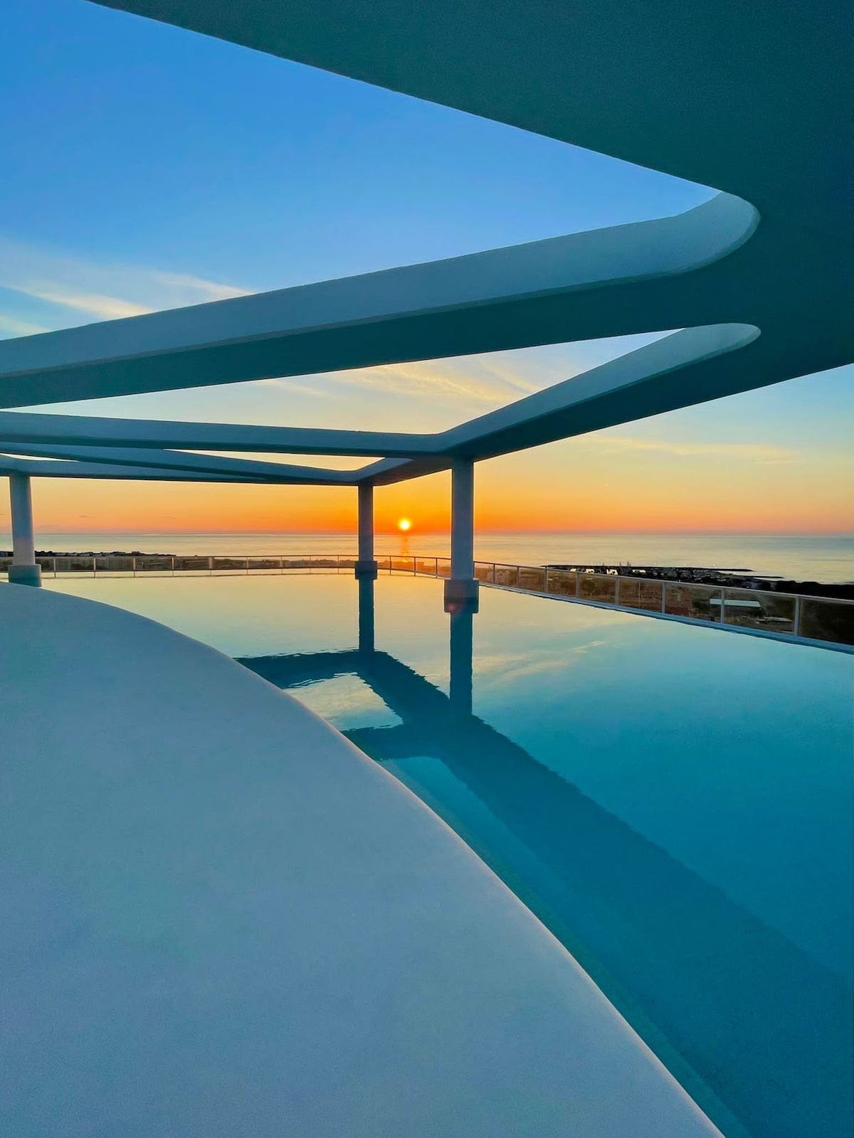 A serene rooftop pool reflects the vibrant colors of a sunset, with smooth white lines of the architecture contrasting against the water's surface. The horizon showcases a peaceful sea, enhancing the tranquil atmosphere of the space.