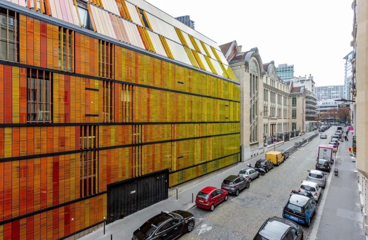 A modern building features a striking facade with colorful panels arranged in horizontal rows, contrasting with an adjacent classic-style structure. The surrounding street is lined with cars parked on cobblestones, creating a mix of contemporary and traditional architectural elements.