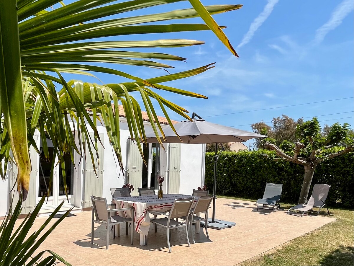 An outdoor terrace is visible, furnished with a dining table set for six, featuring a large umbrella for shade. Two lounge chairs are positioned nearby on a grass lawn. Surrounding greenery creates a private atmosphere, enhancing the spacious area suitable for relaxation.