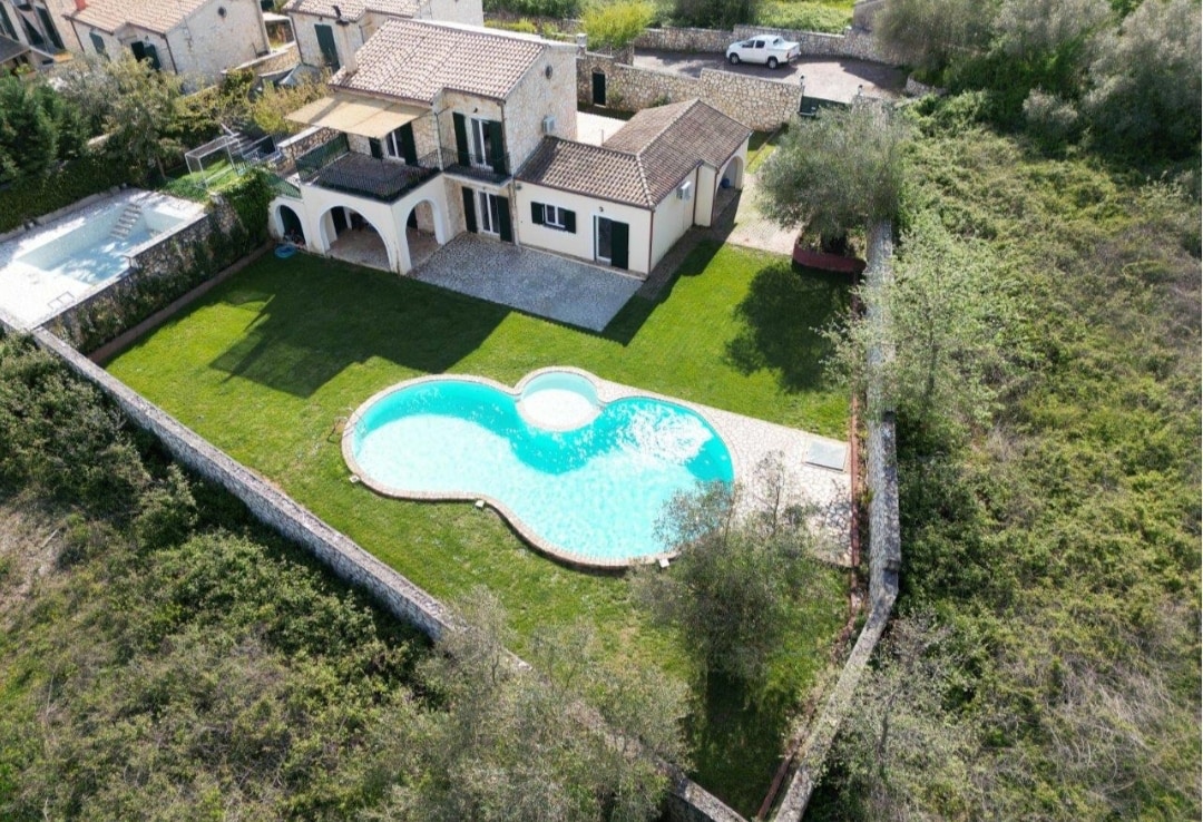 Aerial view of a spacious property featuring a uniquely shaped pool surrounded by lush greenery. The house displays a combination of stone and stucco surfaces with a tiled roof. A well-maintained lawn extends around the pool area, enhancing the outdoor environment.
